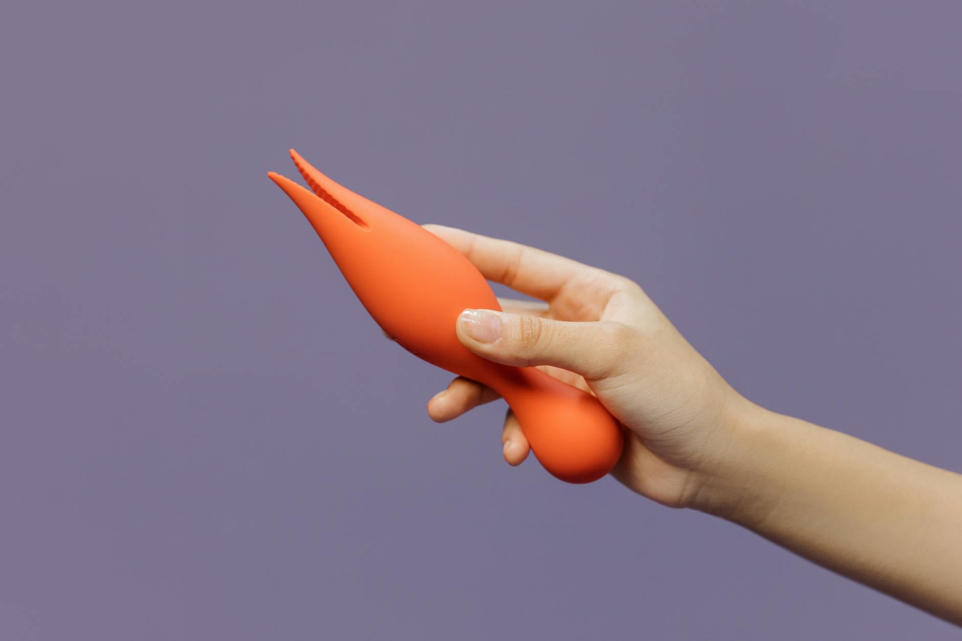 Hand holding a modern orange vibrator against a minimalistic purple backdrop
